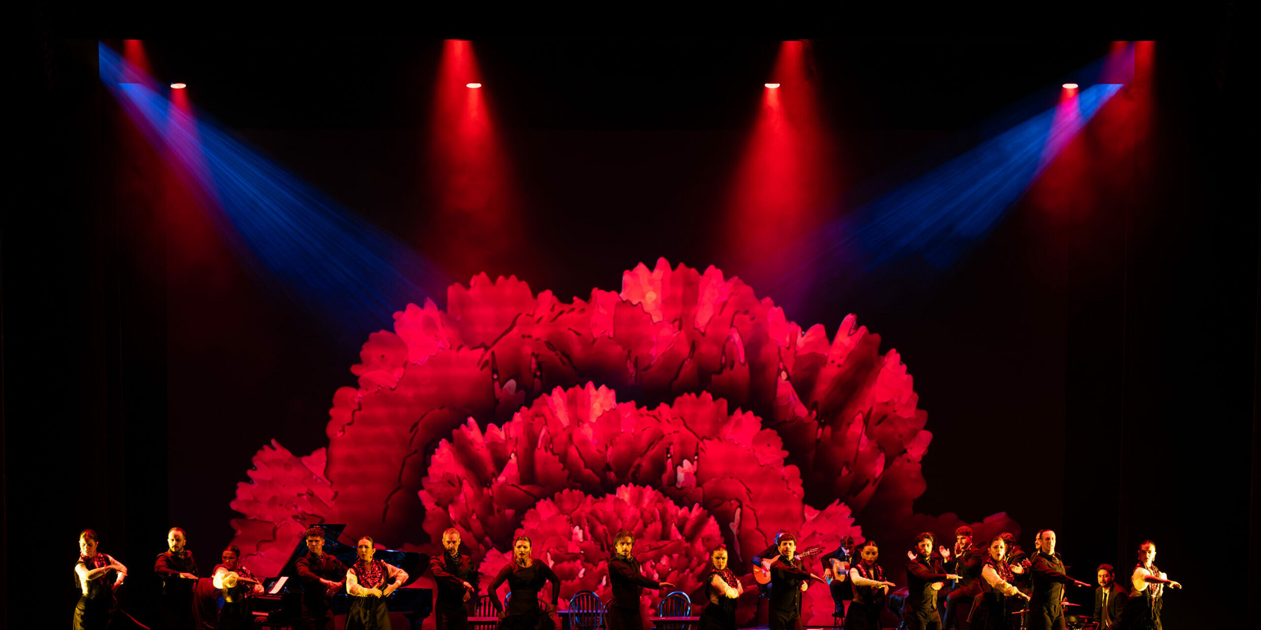 Elenco de 25 artistas de la Compañía Flamenca Antonio Andrade bailando en el escenario de la Royal Opera House de Muscat durante el espectáculo Noches Flamencas, bajo iluminación escénica roja y azul con una gran flor proyectada de fondo.