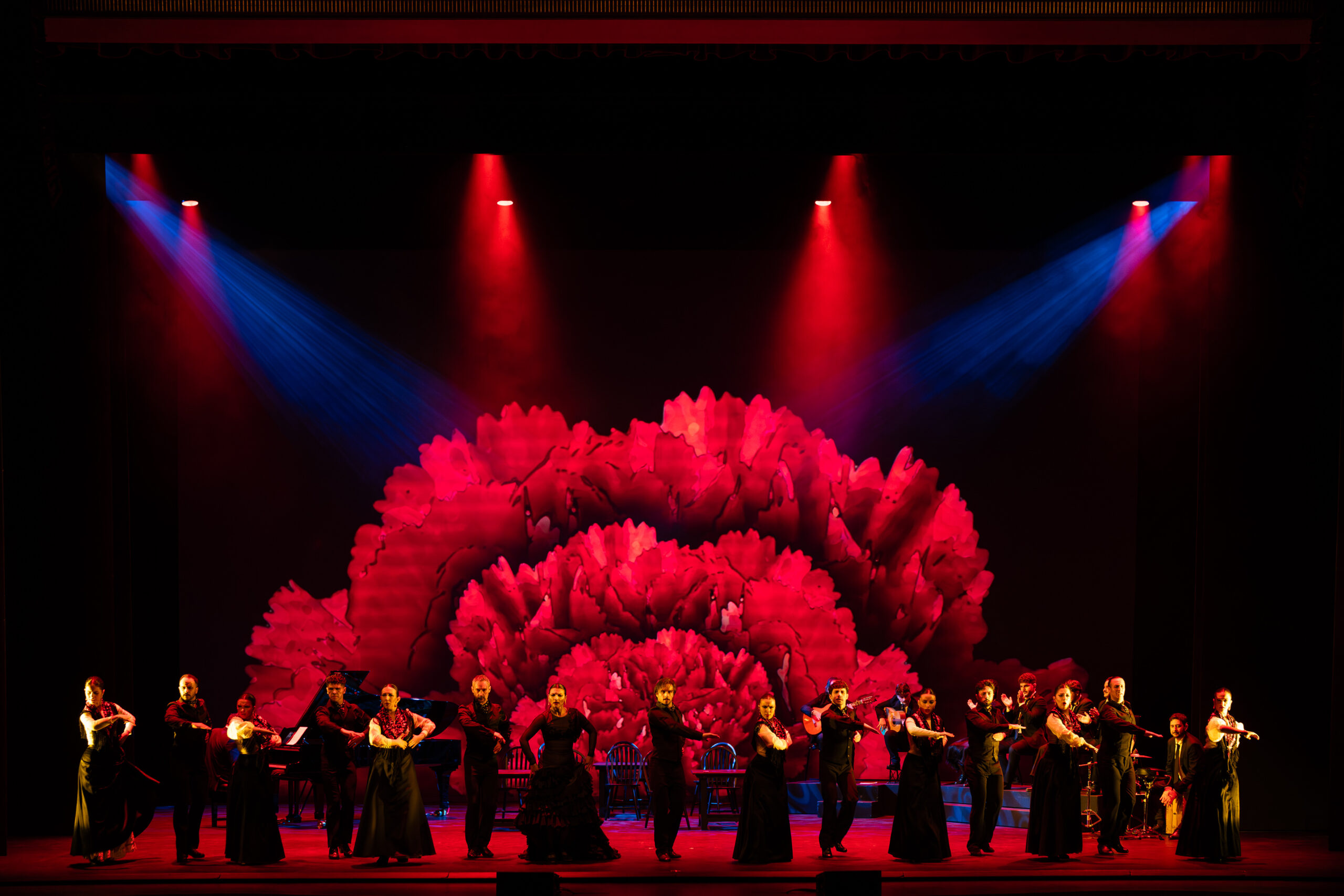 Elenco de 25 artistas de la Compañía Flamenca Antonio Andrade bailando en el escenario de la Royal Opera House de Muscat durante el espectáculo Noches Flamencas, bajo iluminación escénica roja y azul con una gran flor proyectada de fondo.